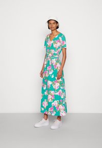Vestido maxi floral em turquesa com flores rosas e brancas, mangas curtas, decote em V, cintura anudada, combinado com ténis brancos e um chapéu de balde branco.