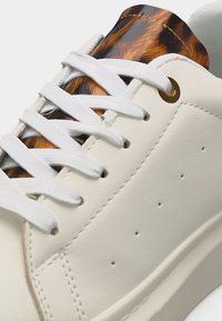 Λευκό αθλητικό παπούτσι με λευκά κορδόνια και καφέ μαύρη μαρμάρινη λεπτομέρεια στη γλώσσα, με εμφανή ραφή και μικρές πλευρικές τρύπες για αερισμό.