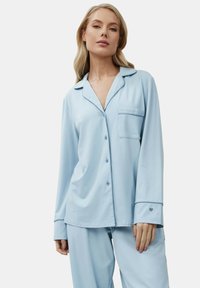 Femme portant un haut de pyjama à manches longues bleu clair avec poche poitrine et un pantalon assorti, sur fond uni.