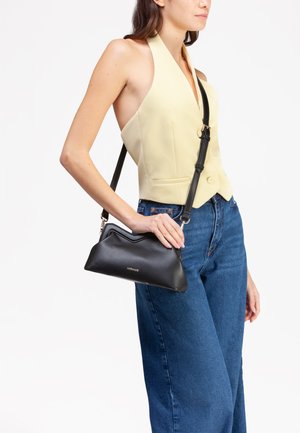 Borsa a tracolla in pelle nera con un design ondulato, dotata di una tracolla removibile e dettagli in hardware dorato, tenuta da una persona che indossa un top giallo.