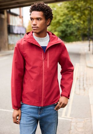 Junger Mann trägt eine rote Kapuzenjacke und blaue Jeans und steht in einer städtischen Straße mit Grün im Hintergrund.