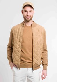 Chaqueta bomber acolchada color beige con cuello de canalé, llevada sobre un suéter de punto a juego. La modelo también lleva una gorra beige y pantalones blancos.