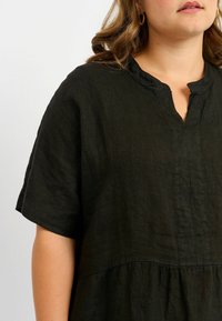 Schwarzes Leinenoberteil mit lockerer Passform, kurzen Ärmeln und V-Ausschnitt. Der strukturierte Stoff verfügt über eine geraffte Naht in der Taille.