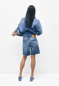 Chaqueta de mezclilla con mangas de gran tamaño, combinada con pantalones cortos de mezclilla a juego. Presenta una etiqueta naranja visible en la parte posterior y un dobladillo deshilachado. Color azul claro.