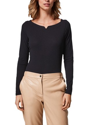 Kvinde iført en sort langærmet top med en let V-udskæring og beige højttaljede bukser med knaplukning.