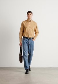 Mann som går, iført en lys brun skjorte med knapper, blå jeans, svart belte og sko, holder en brun skulderveske mot en ensfarget lys bakgrunn.