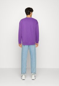 Sudadera morada de gran tamaño con puños y dobladillo acanalados, combinada con jeans de mezclilla azul claro y zapatillas blancas. Presenta un diseño sencillo.