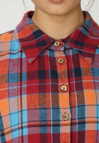 Chemise à carreaux avec des couleurs rouge, bleu et orange. Elle possède un col pointu et des boutons en bois le long de la patte de boutonnage. Texture de tissu douce.