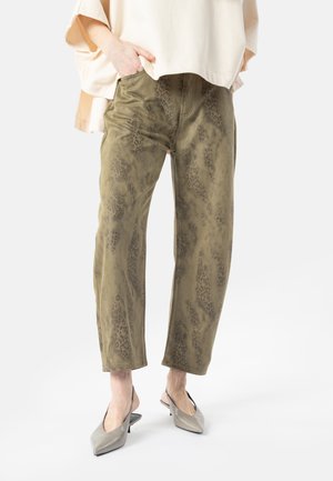 Person iført olivengrønne leopardprintede cropped bukser med beige oversized top og grå spidse slingback-hæle, stående med hænderne i lommerne.