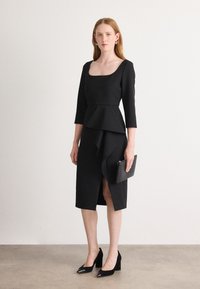 Abito peplum nero con maniche a tre quarti, orlo asimmetrico e spacco laterale. Indossato con tacchi a punta e una clutch nera. Texture liscia.