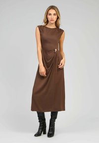 Robe sans manches marron avec un design drapé, présentant un nœud sur le côté et un accent circulaire. Associée à des bottes noires montantes. Texture lisse.