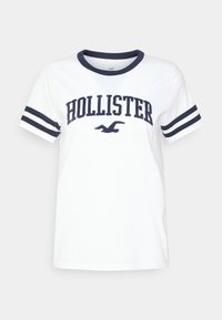 White t-shirt with navy blue collar and sleeve stripes. Features "HOLLISTER" in bold, navy font across the chest. Soft cotton material.
