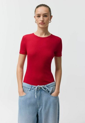 Kobieta z włosami związanymi w kok, ubrana w czerwoną bluzkę z krótkim rękawem i luźne jasnoniebieskie dżinsy ze sznurkiem, ręce w kieszeniach, neutralne tło.