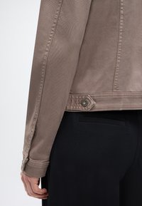 Άτομο με ταμπά κοντό σακάκι με λεπτομέρειες κουμπιών πάνω από μαύρο παντελόνι ραμμένο, εμφανίζοντας το χέρι και την πίσω πλευρά.