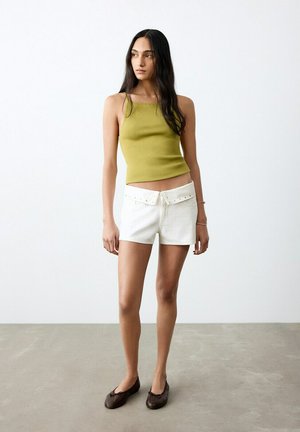 Donna in piedi che indossa una canotta verde oliva senza maniche, pantaloncini bianchi con borchie e vita risvoltata, e ballerine marrone scuro.