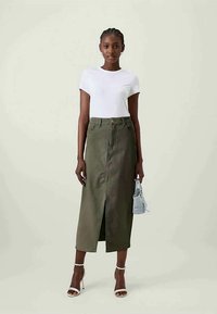 Saia de ganga verde-oliva com fenda frontal, combinada com uma t-shirt branca simples. Modelo a usar sapatos de salto abertos brancos e a segurar uma pequena mala azul.