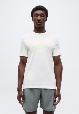 Άνδρας που στέκεται, φοράει λευκό μπλουζάκι Nike με υπόξανθο και πράσινο λογότυπο και γκρι αθλητική βερμούδα, ουδέτερο φόντο.