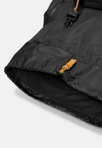 Giacca imbottita nera con una superficie liscia, dotata di chiusura a zip e un dettagli in pelle marrone chiaro sul fondo. Design semplice e funzionale.
