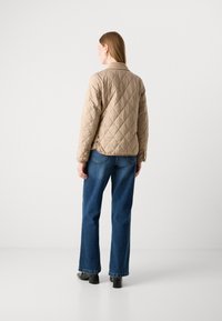 Kvindelig i en beige quiltet jakke, blå lige ben jeans og sorte hæle, står med ryggen til mod en ensfarvet hvid baggrund.