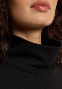 Close-up van iemands ondergezich met glanzende lippen, gekleed in een zwart hooggegesloten kledingstuk, en met een deel van het donkere, krullende haar zichtbaar.