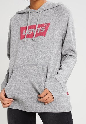 Personne portant un sweat à capuche Levi's gris clair avec une poche avant et un logo rouge sur la poitrine, associé à un pantalon noir.