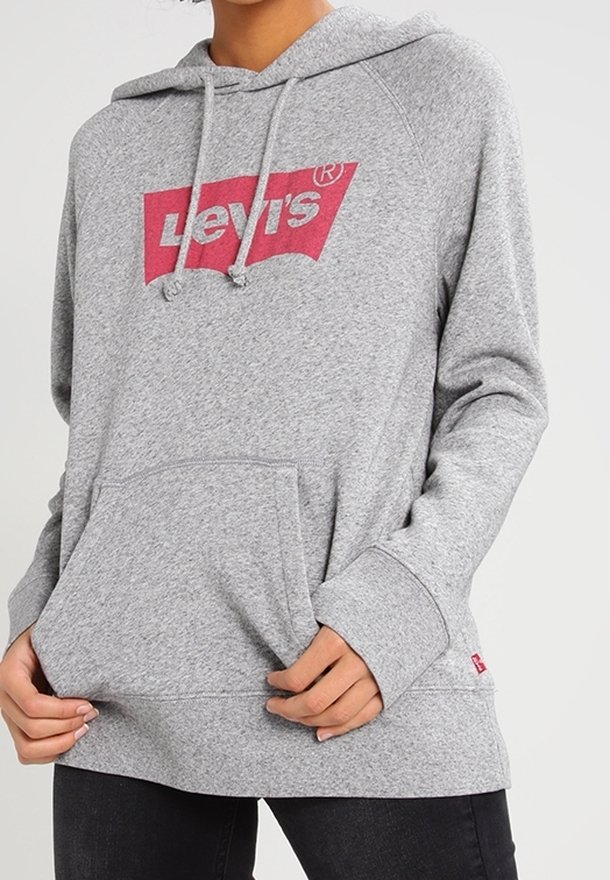 Personne portant un sweat à capuche Levi's gris clair avec une poche avant et un logo rouge sur la poitrine, associé à un pantalon noir.