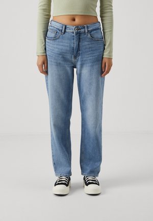 Personne portant un jean droit bleu clair, des baskets noires et blanches, et un crop top vert clair à manches longues, debout devant un fond blanc.