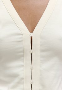 Cremfarbendes Hemd mit V-Ausschnitt, strukturiertem Stoff und fünf Knöpfen auf der Vorderseite, mit sichtbarem Knopfverschluss.