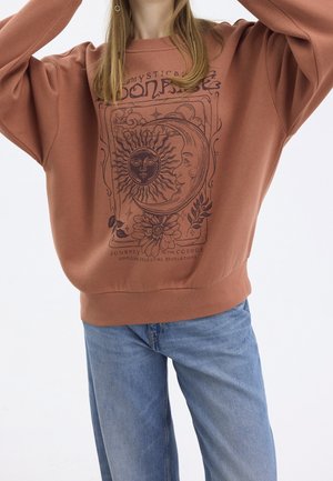 Femme portant un sweat-shirt couleur rouille avec un motif céleste de soleil, lune et fleur, associé à un jean bleu clair, sur fond blanc uni.