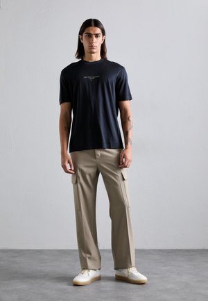 Tânăr cu păr lung și întunecat, îmbrăcat cu un tricou negru cu logo, pantaloni cargo bej și adidași albi, stând în fața unui perete simplu de culoare deschisă.