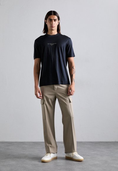 Jonge man met lang, donker haar, gekleed in een zwart t-shirt met logo, beige cargo pants en witte sneakers, staand tegen een effen lichte muur.