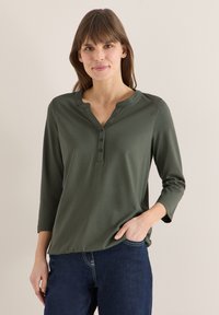 Femme aux cheveux bruns lisses portant un haut henley à manches longues vert olive et un jean bleu foncé, debout avec une main dans la poche.