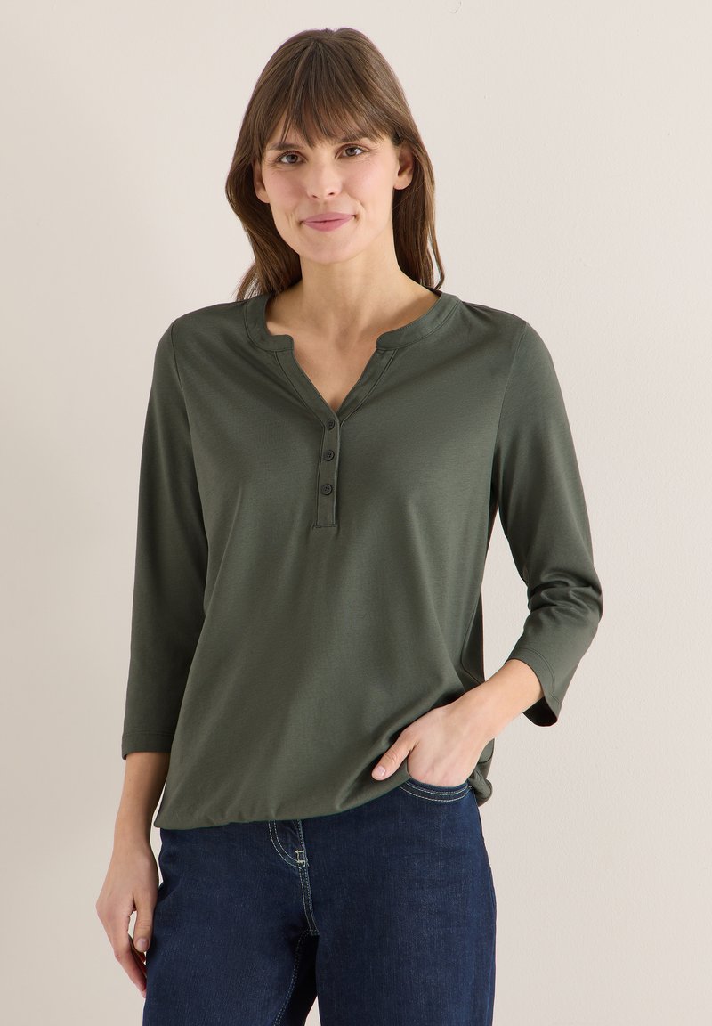 Femme aux cheveux bruns lisses portant un haut henley à manches longues vert olive et un jean bleu foncé, debout avec une main dans la poche.