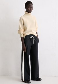 Wysoka osoba o ciemnej karnacji, ubrana w kremowy sweter z golfem i luźne czarne spodnie z białymi paskami po bokach, stojąca przy gładkiej ścianie.