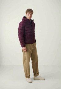 Bordeaux puffy jakke med horisontal syning, lynlås foran og hætte. Parret med khaki bukser og lyse sneakers. Minimalistisk detaljering.