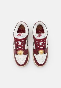 Nike sneakers feature maroon leather accents, white perforated leather, red laces, and a gold emblem on the tongue. Classic design and round toe shape.