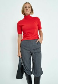 Top rojo de manga corta con cuello alto combinado con pantalones grises de corte a medida. La modelo sostiene un bolso negro y lleva botas negras hasta la rodilla.
