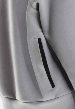 Pelēka sporta kreklu audums ar gludu tekstūru; tam ir melna ar rāvējslēdzēju aizverama kabata kreisajā piedurknes pusē, lai nodrošinātu papildu funkcionalitāti.