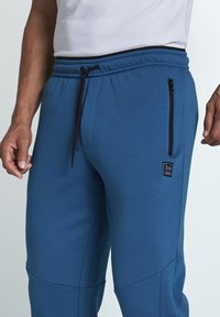 Blaue Sweatpants aus weichem Stoff mit einem elastischen Bund, verstellbarem Kordelzug und seitlichen Reißverschlusstaschen. Kleine Logodetails am Bein.