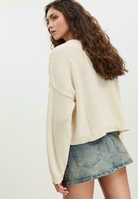 Vrouw met golvend bruin haar draagt een crèmekleurige gebreide trui en een versleten denim minirok, staand tegen een effen lichte achtergrond.