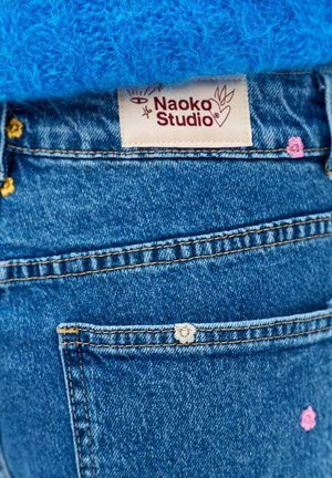 Kieszeń dżinsów z małymi haftowanymi kwiatami i metką Naoko Studio powyżej, zestawiona z niebieską dzianinową tkaniną.