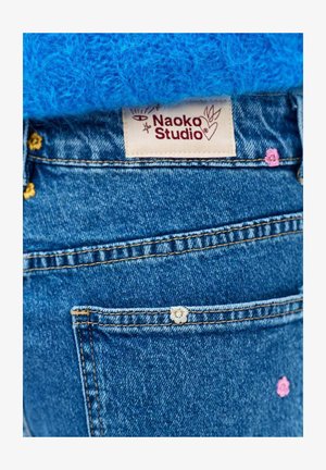 Kieszeń dżinsów z małymi haftowanymi kwiatami i metką Naoko Studio powyżej, zestawiona z niebieską dzianinową tkaniną.