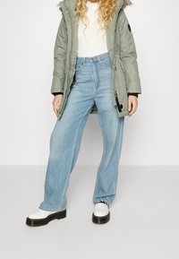 Parka grigia con bordo in pelliccia e cerniera, maglietta bianca a maniche lunghe, jeans a gamba larga blu chiaro e scarpe bianche con lacci e suola nera.