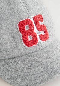 Grå baseballcap i filt med hævet rødt broderet nummer "85" indrammet i hvidt, med en tekstureret overflade og buet skygge.
