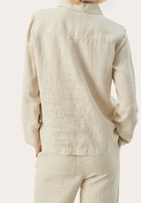 Person i en lys beige linnedragt og matchende bukser, vist fra ryggen med hænderne i lommerne, mod en ensfarvet baggrund.