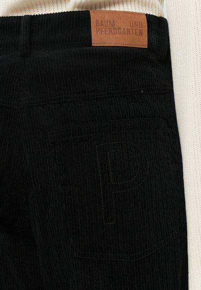Melna korduroja bikses ar vertikālām ribām, ar brūnu ādas jostas etiķeti un izšūtu burtiem "P" aizmugurējā kabatā.