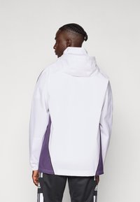 Hvid vandtæt jakke med et lilla sidepanel, justerbar hætte og elastiske manchetter. Glat tekstur med minimal synlig branding.