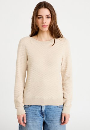 Beige trui met een gestructureerd honingraatpatroon. Voorzien van een ronde halslijn en geribbelde manchetten. Gecombineerd met lichtblauwe spijkerbroek.