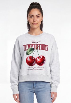 Jeune femme portant un sweat-shirt gris clair avec de grosses cerises rouges et le texte « Sweet Temptations », debout sur un fond blanc.