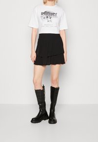 Žena oblečená v bílé grafické zkrácené tričko, černé vrstvené mini sukně a černých vysokých robustních botách stojí na jednobarevném pozadí.